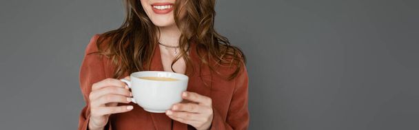 cropped view of happy young woman with brunette hair wearing brown and trendy suit with blazer and holding cup of coffee on grey background, work-life balance, banner  - Photo, Image