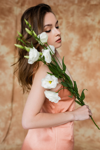 portrait of brunette and young woman in pink silk slip dress holding eustoma flowers while standing on mottled beige background, sensuality, elegance, sophistication  - Fotoğraf, Görsel