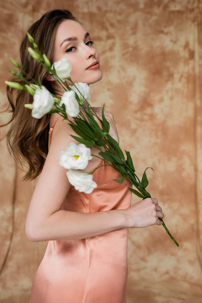 portrait de gracieuse, brune et jeune femme en soie rose slip robe tenant des fleurs d'eustomie tout en se tenant debout sur fond beige moucheté, sensualité, élégance, sophistication, regardant la caméra  - Photo, image