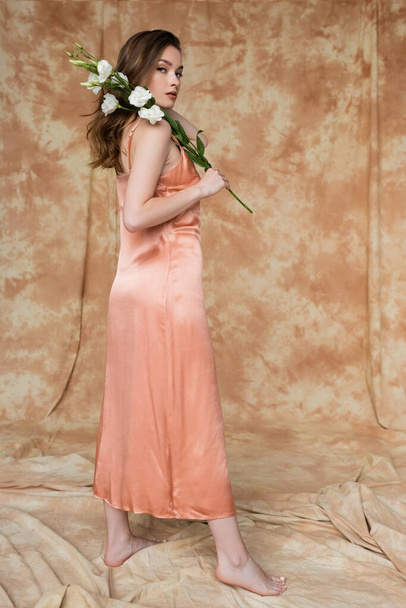 πλήρες μήκος του ξυπόλυτη και μελαχρινή και νεαρή γυναίκα σε ροζ μεταξωτό φόρεμα ολίσθησης κρατώντας λευκά λουλούδια, ενώ στέκεται σε στίγματα μπεζ φόντο, αισθησιασμό, κομψότητα, κομψότητα  - Φωτογραφία, εικόνα