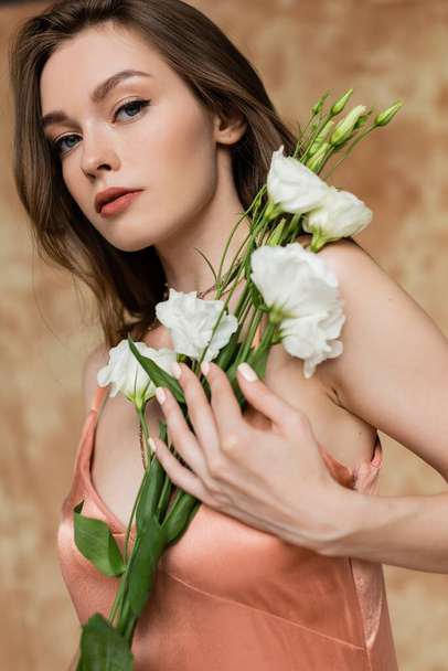 portrait of captivating and young woman in pink silk slip dress holding eustoma flowers while standing and looking at camera on mottled beige background, sensuality, elegance, sophistication  - Foto, Imagem