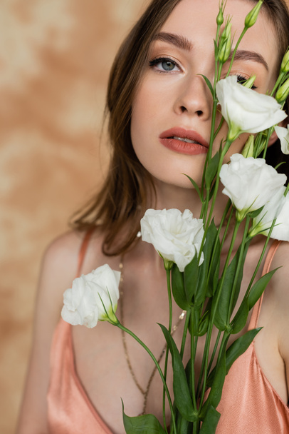 portrait of sensual and young woman holding eustoma flowers while standing and looking at camera on mottled beige background, captivating beauty, elegance, sophistication  - Foto, Bild