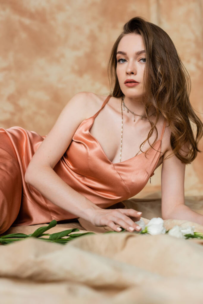 pretty young woman in pink silk slip dress lying on blurred linen fabric and looking at camera near white flowers on mottled beige background, sensuality, sophistication, elegance, eustoma  - Foto, imagen