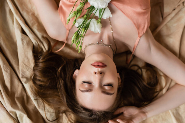 Draufsicht der verführerischen jungen Frau im rosa Seidenkleid mit geschlossenen Augen auf Leinenstoff mit zarten weißen Blüten auf beigem Hintergrund liegend, Sinnlichkeit, verkehrte Sicht  - Foto, Bild