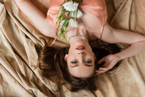 πάνω όψη σαγηνευτική νεαρή γυναίκα σε ροζ μεταξωτό ολίσθημα φόρεμα ξαπλωμένη σε λινό ύφασμα με λεπτά λευκά λουλούδια και κοιτάζοντας κάμερα σε μπεζ φόντο, αισθησιασμό, αναποδογυρισμένη άποψη  - Φωτογραφία, εικόνα