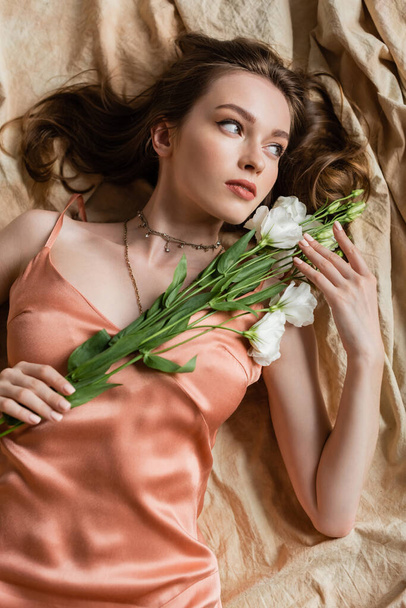 top view of attractive young woman in pink silk slip dress lying on linen fabric and holding delicate white flowers on beige background, sensuality, elegance, eustoma, looking away  - Fotografie, Obrázek