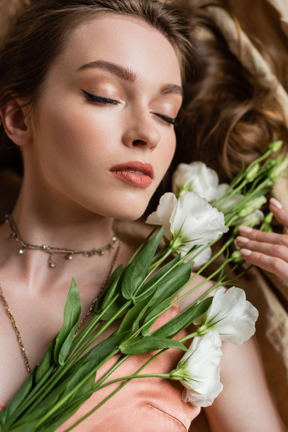 top view of attractive young woman in pink silk slip dress lying with closed eyes on linen fabric and holding white flowers on beige background, sensuality, elegance, eustoma, portrait  - 写真・画像