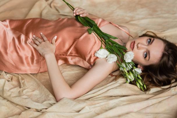 atractiva mujer joven en vestido de seda rosa deslizamiento acostado en tela de lino y la celebración de delicadas flores blancas sobre fondo beige moteado, sensualidad, sofisticación, elegancia, eustoma, mirando a la cámara  - Foto, imagen