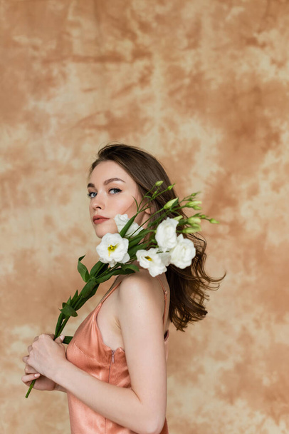 young woman with brunette hair posing in pink and silk slip dress and holding white eustoma flowers on mottled beige background, sensuality, sophistication, elegance, looking at camera - Фото, зображення