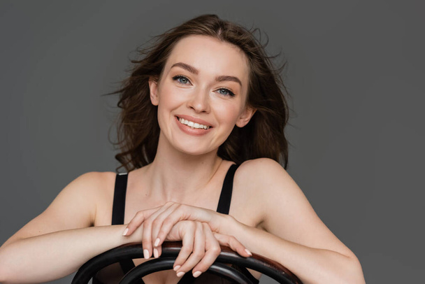portrait of beautiful and happy young woman with brunette hair and blue eyes smiling while leaning on chair back and looking at camera isolated on grey background, sensuality, grace, feminine beauty  - Photo, Image