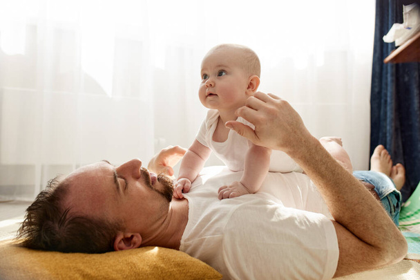 Happy dad holds his newborn baby in his arms while lying on the floor in a bright childs room. The father looks at the child with a loving look. Happy childhood and fatherhood - Foto, Bild