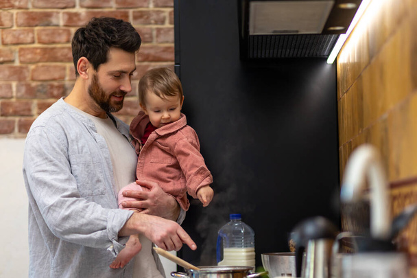 Bearded man cooking dinner with baby daughter in hands standing in kitchenn, father with infant baby. - Foto, afbeelding