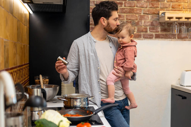 Barbudo hombre cocina cena con bebé hija en manos de pie en kitchenn, padre besando bebé bebé. - Foto, imagen