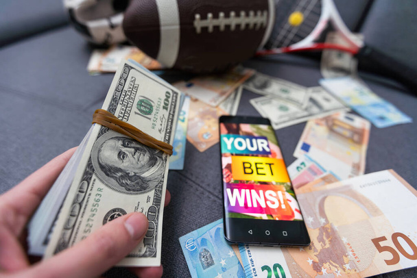 Online sportweddenschappen. Dollars vallen op de achtergrond van een hand met een smartphone en een voetbal. Creatieve achtergrond, gokken - Foto, afbeelding
