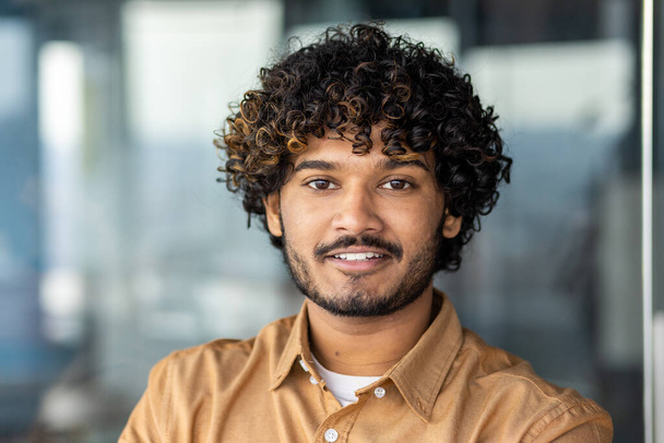 Молодой улыбающийся индийский программист крупным планом улыбается и смотрит в камеру, портрет человека с вьющимися волосами и рубашкой внутри офиса на работе, бизнесмен-предприниматель с бородой работает над проектом - Фото, изображение