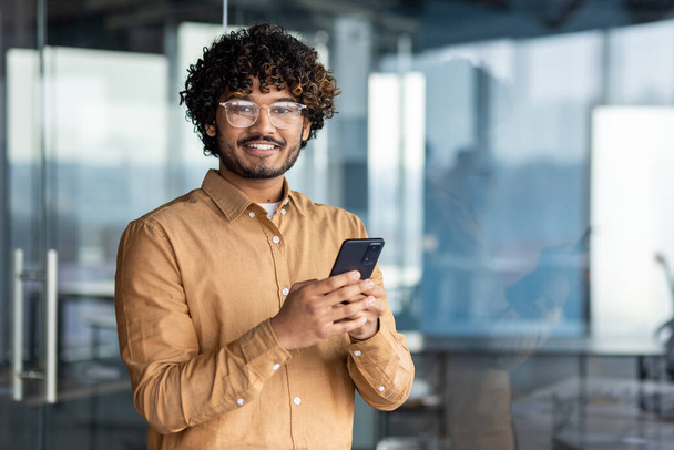 Portrait of successful businessman inside modern bright office, indian man smiling and looking at camera, man in shirt and glasses holding phone. - Foto, imagen