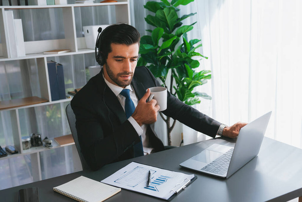 Manager of call center operator office sitting on his desk with his coffee while working on laptop. Office worker wearing headset and black suit working on customer support or telemarketing. fervent - Foto, imagen