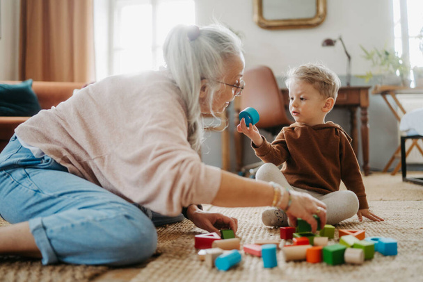 Grandmother playing with her little grandson, building a wooden block set in his room. - Foto, Bild