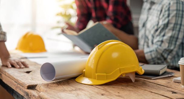 Yellow hard hat on table and house design print design with construction team planning start new project plan in office at building site partner and contractor. - Φωτογραφία, εικόνα