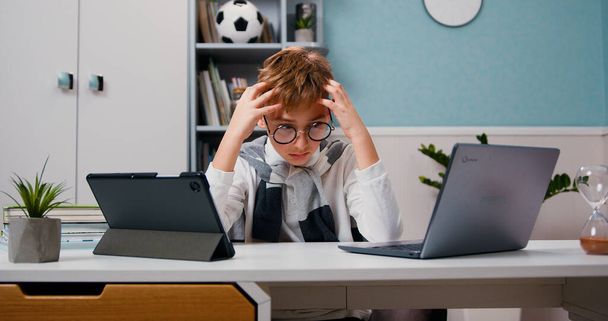 Teini surullinen poika istuu kyllästyneenä kotona kannettavan tietokoneen ja tabletin kanssa etäopetuksen aikana ja pitää päänsä. - Valokuva, kuva