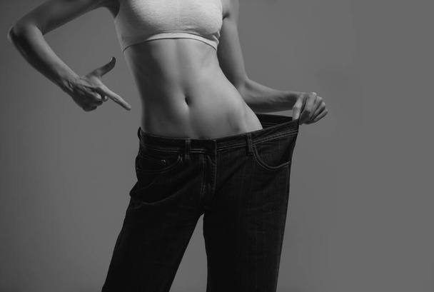 Dieting concept with oversized jeans. Skinny woman in too large jeans. Concept of successful weight loss. Thin girl in big trousers on a gray background - Zdjęcie, obraz