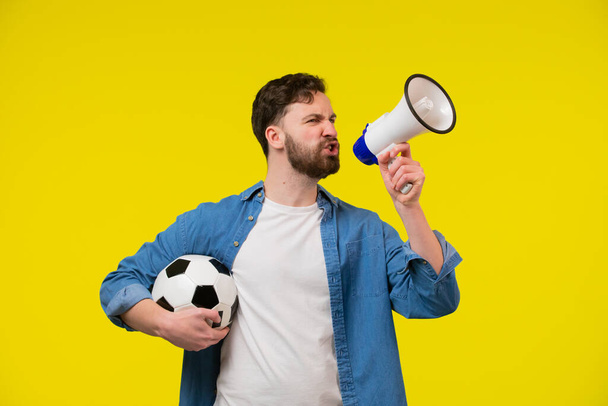Very happy cheerful man with beard in striped t-shirt blowing in big bullhorn holding football ball, celebrating championship beginning. Indoor studio shot isolated on yellow background - Fotó, kép