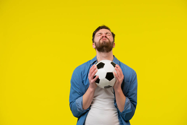 Возбужденный молодой бородатый футбольный фанат в белой футболке поддерживает любимую команду с футбольным мячом, кричащим сжимая кулак изолированным на желтом фоне. Семейный досуг. - Фото, изображение