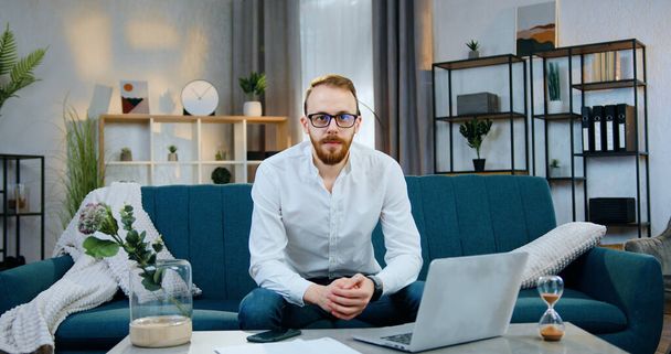 Handsome confident in good mood young bearded man in glasses sitting on comfortable sofa at home and looking into camera ,front view - 写真・画像