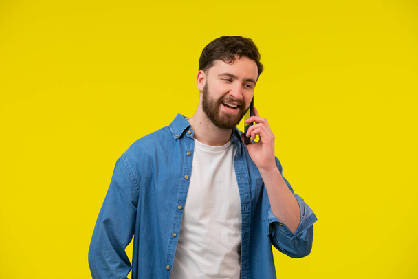 Young smiling happy cheerful caucasian man 20s wear white casual shirt talk speak on mobile cell phone conducting pleasant conversation look aside isolated on plain yellow background studio portrait. - Фото, изображение