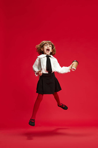 Full-length portrait of happy, smiling, little girl in black shirt and white blouse jumping with coffee and tablet against red studio background. Concept of childhood, education, fashion, kid emotions - Photo, Image