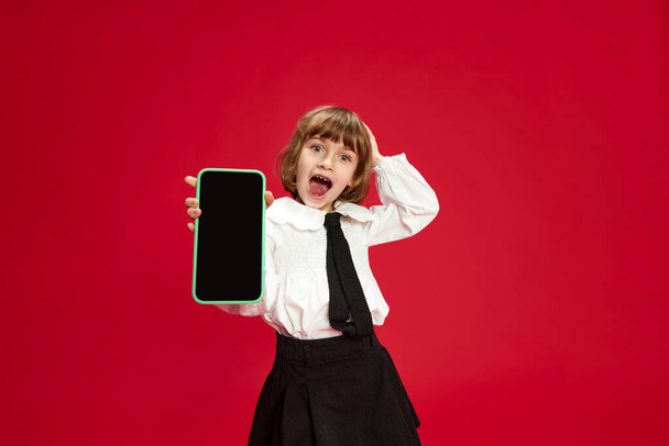 Retrato de niña feliz, sonriente y riente en blusa blanca y camisa negra, mostrando la pantalla del teléfono móvil contra el fondo rojo del estudio. Concepto de infancia, educación, moda, emociones infantiles - Foto, Imagen