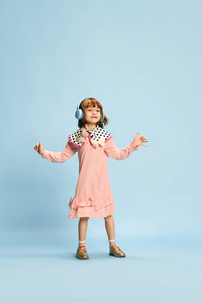 Ganztägiges Porträt eines entzückenden, glücklichen, lächelnden kleinen Mädchens in niedlichem rosa Kleid, das mit Kopfhörern auf blauem Studiohintergrund Musik hört. Konzept von Kindheit, Emotionen, Spaß, Mode, Lebensstil - Foto, Bild