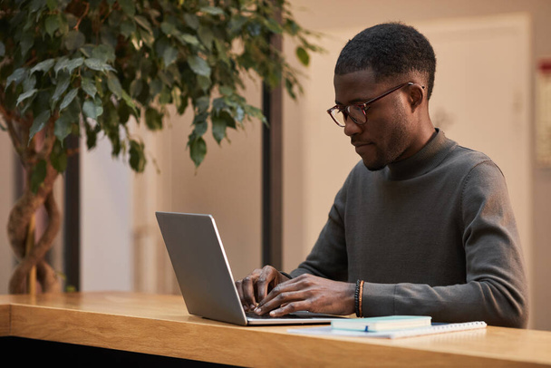 現代オフィスのデスクでラップトップを使用して若い黒人ビジネスマンのミニマルな暖かいトーンの肖像画、コピースペース - 写真・画像