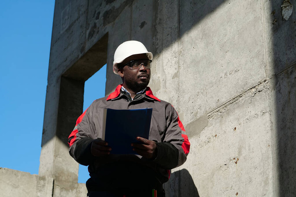 Afrykański amerykański robotnik stoi na placu budowy z dokumentami roboczymi - Zdjęcie, obraz