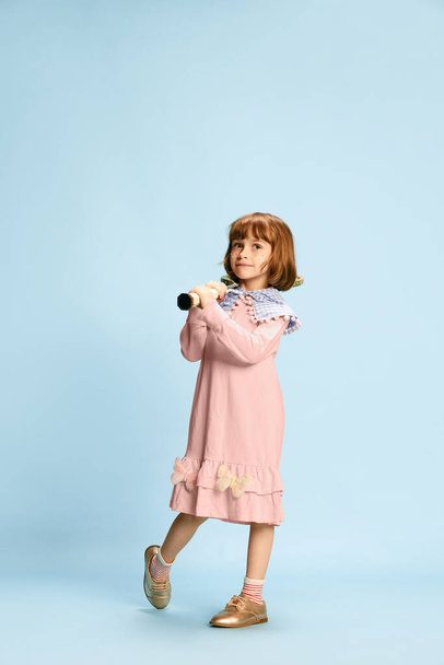 Full-length portrait of smiling, happy, little girl in pink dress posing with tennis racket against blue studio background. Concept of childhood, emotions, fun, fashion, active lifestyle, sport - Foto, afbeelding
