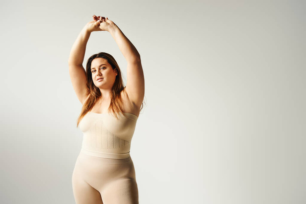 plus size woman with natural makeup posing with raised hands in beige strapless top and underwear in studio isolated on grey background, looking at camera, body positive, self-confidence   - Photo, Image