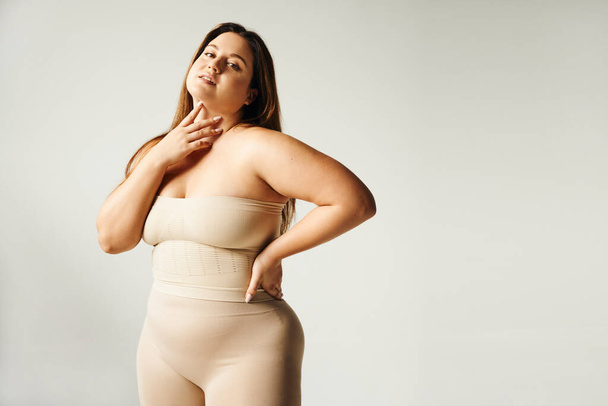 Woman with plus size body touching her neck and looking at camera while posing with hand on hip in beige strapless top and underwear in studio isolated on grey background, body positive, self-love  - Photo, Image