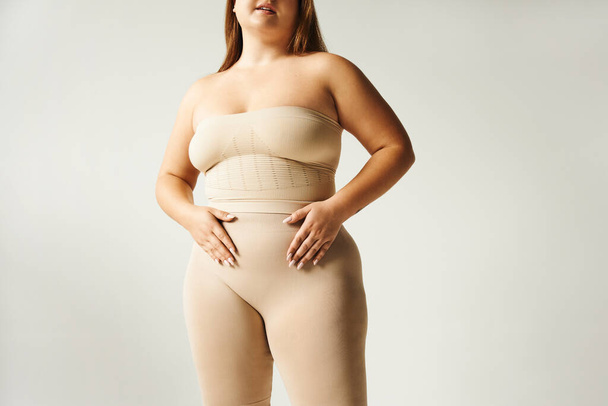 bijgesneden beeld van vrouw met plus size lichaam poseren in strapless top met blote schouders en ondergoed geïsoleerd op grijze achtergrond in studio, lichaam positief, zelf-liefde, handen op de buik  - Foto, afbeelding