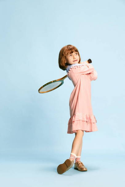 Full-length portrait of smiling, cute, little girl in pink dress posing with tennis racket against blue studio background. Concept of childhood, emotions, fun, fashion, active lifestyle, sport - Photo, Image