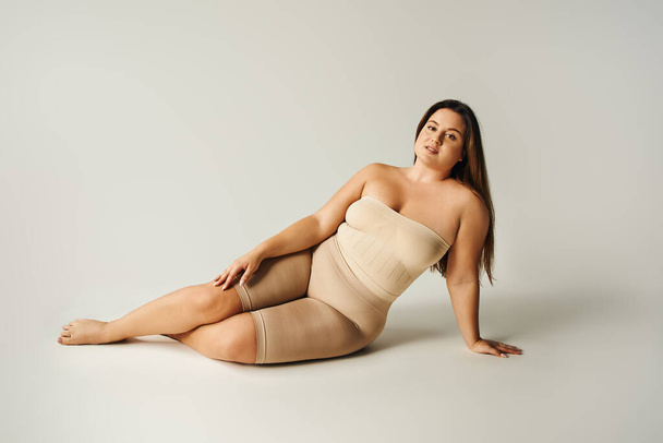 barefoot and confident woman in strapless top with bare shoulders and underwear posing while sitting in studio on grey background, body positive, self-love, plus size  - Foto, imagen