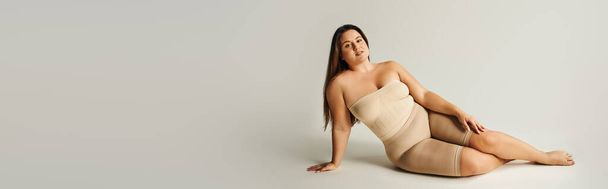 barefoot and confident woman in strapless top with bare shoulders and underwear posing while sitting in studio on grey background, body positive, self-love, plus size, banner  - Photo, Image