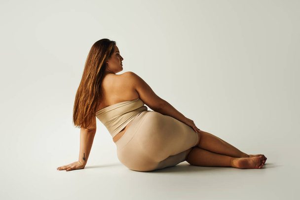 back view of barefoot woman with plus size body in strapless top with bare shoulders and underwear posing while sitting in studio on grey background, body positive, tattoo translation: harmony  - Foto, Bild