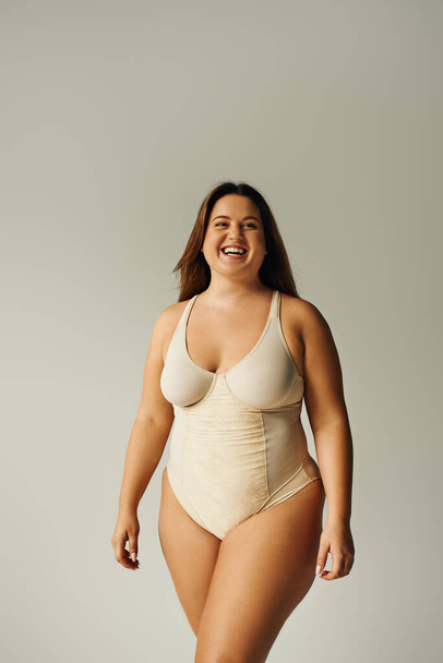 happy plus size woman in beige bodysuit posing while standing in studio on grey background, body positive, figure type, self-esteem, smiling while looking at camera  - Foto, Bild