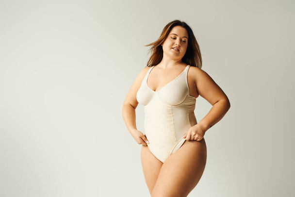 tattooed and brunette curvy woman with plus size body posing in beige bodysuit while standing in studio on grey background, body positive, figure type, self-esteem, tattoo translation: harmony  - Photo, Image