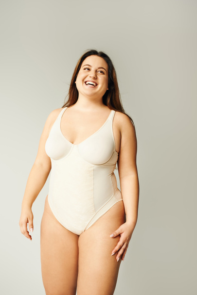 portrait of positive and curvy woman with plus size body posing in beige bodysuit while laughing on grey background, body positive, figure type, looking away while standing in studio  - Foto, imagen
