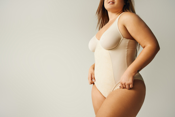 partial view of curvy woman wearing beige bodysuit and standing with hand on hip isolated on grey background, self-confidence, figure type, body positivity movement, tattoo translation: harmony  - Photo, Image