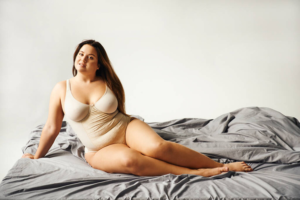 elbűvölő nő természetes smink és plusz méretű test visel bézs testruha és pózol az ágyban szürke ágynemű, test pozitív, figura típusú, csupasz láb, nézi a kamera  - Fotó, kép