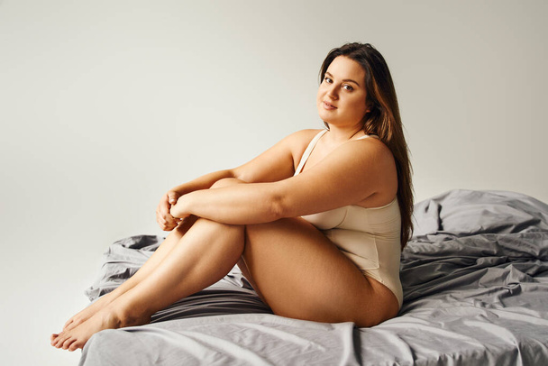 full length of brunette alluring woman with long hair and plus size body and bare feet wearing beige bodysuit and looking at camera while sitting on bed with grey bedding, body positive, figure type  - Фото, изображение