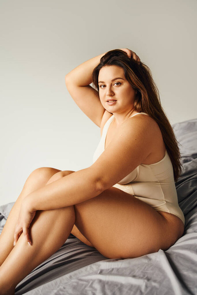 brunette alluring woman with long hair and plus size body and bare feet wearing beige bodysuit and looking at camera while touching hair and sitting on bed with grey bedding, body positive  - Photo, Image