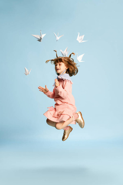 Full-length portrait of playful, lovely little girl in pink dress posing, playing with paper birds against blue studio background. Concept of childhood, emotions, fun, fashion, lifestyle, imagination - Фото, зображення